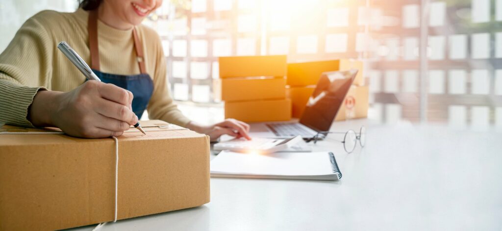 A woman writes on a brown shipping package with a pen, with a laptop, notebook, glasses, and more packages are on the table