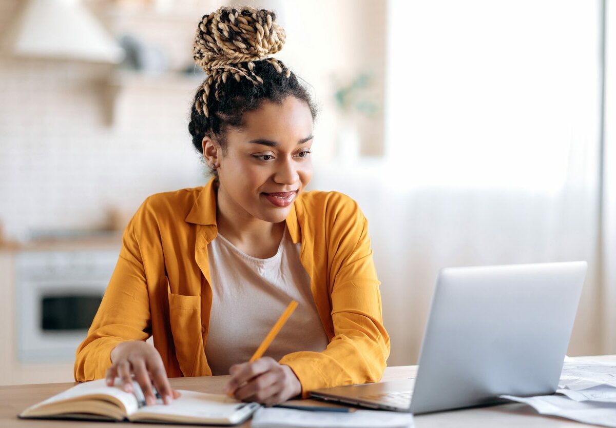 A woman works at a table with her laptop and her home kitchen in the background