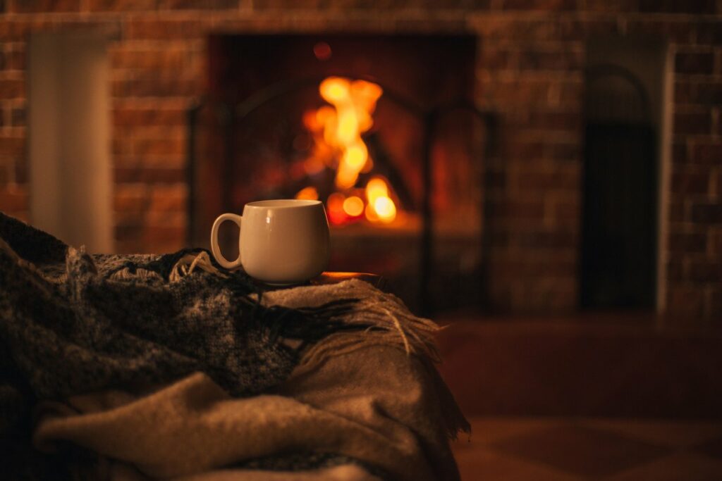 A white mug sits on a small table with blankets and a lit, cozy fireplace nearby