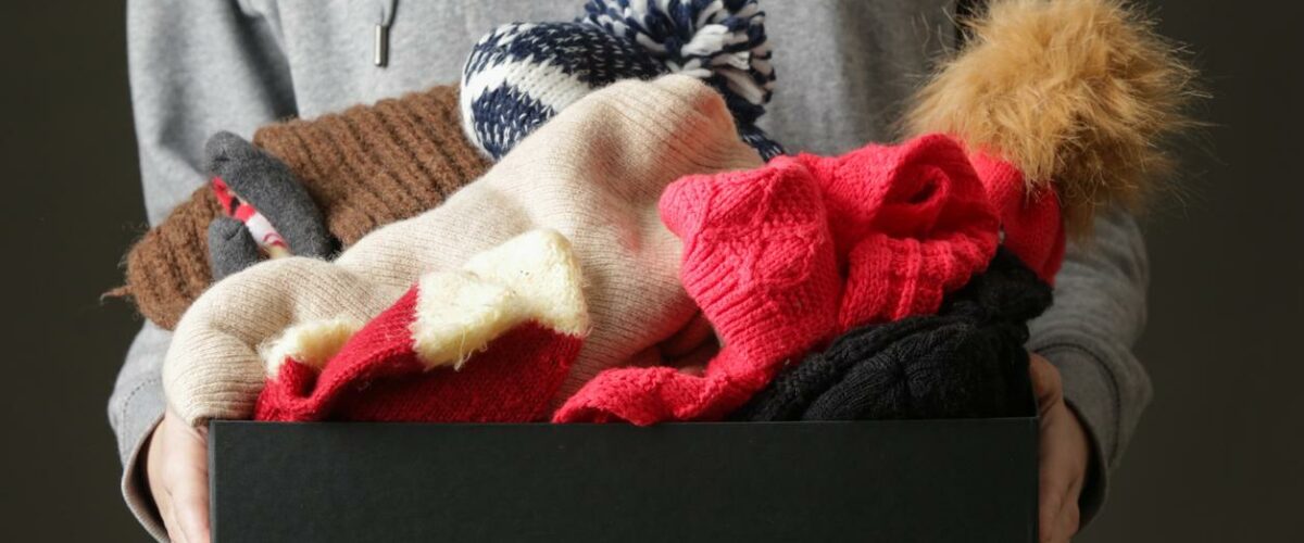A person holds a small box full of winter clothing including hats and gloves
