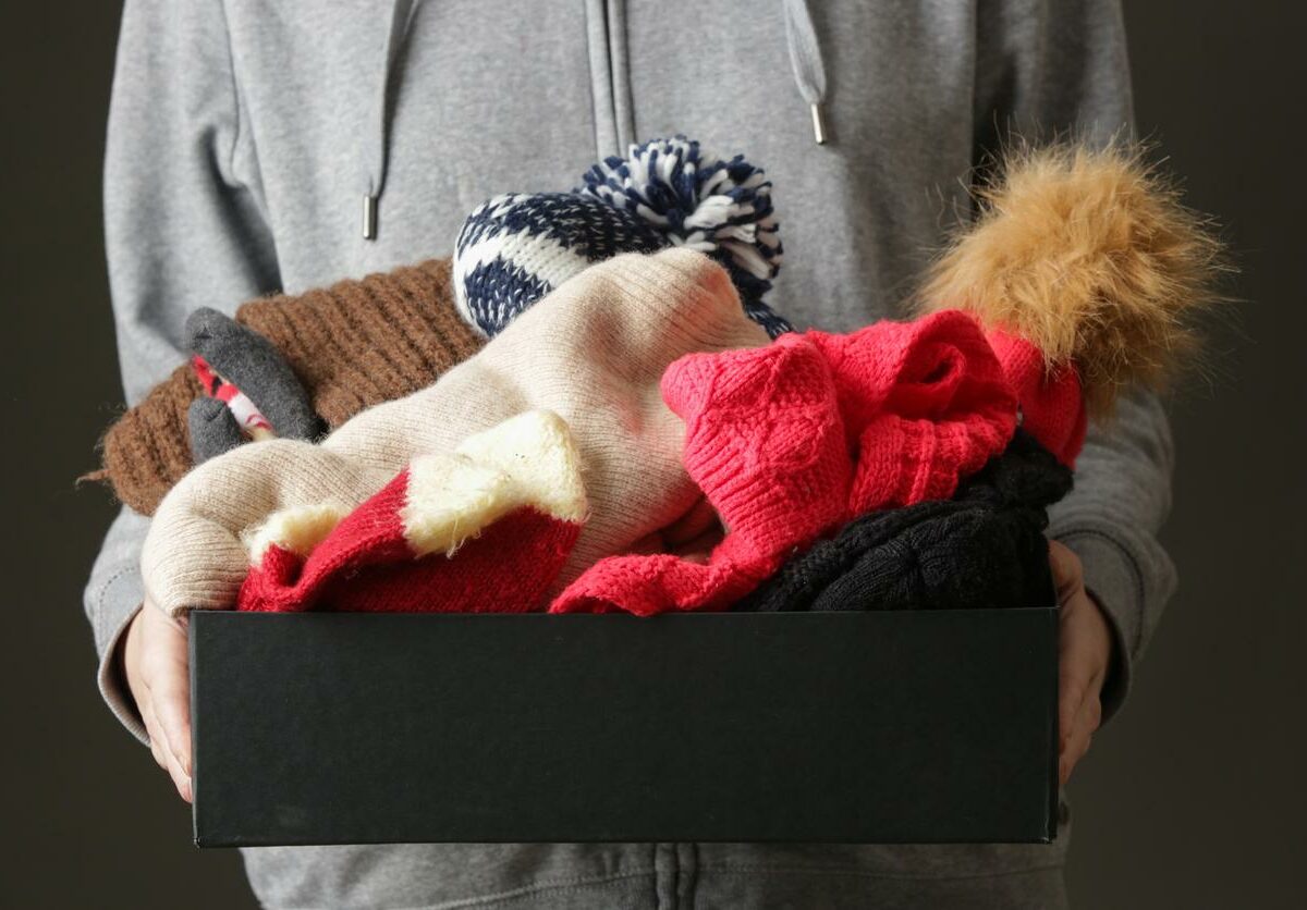 A person holds a small box full of winter clothing including hats and gloves