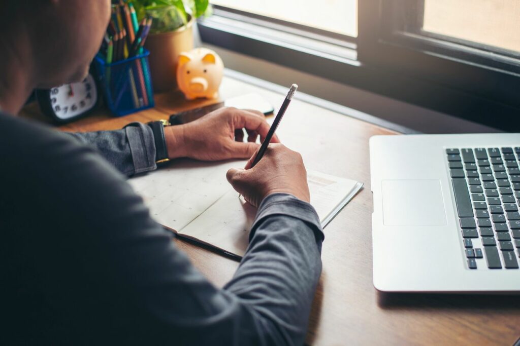 A person writes notes in their planner with an open laptop beside them