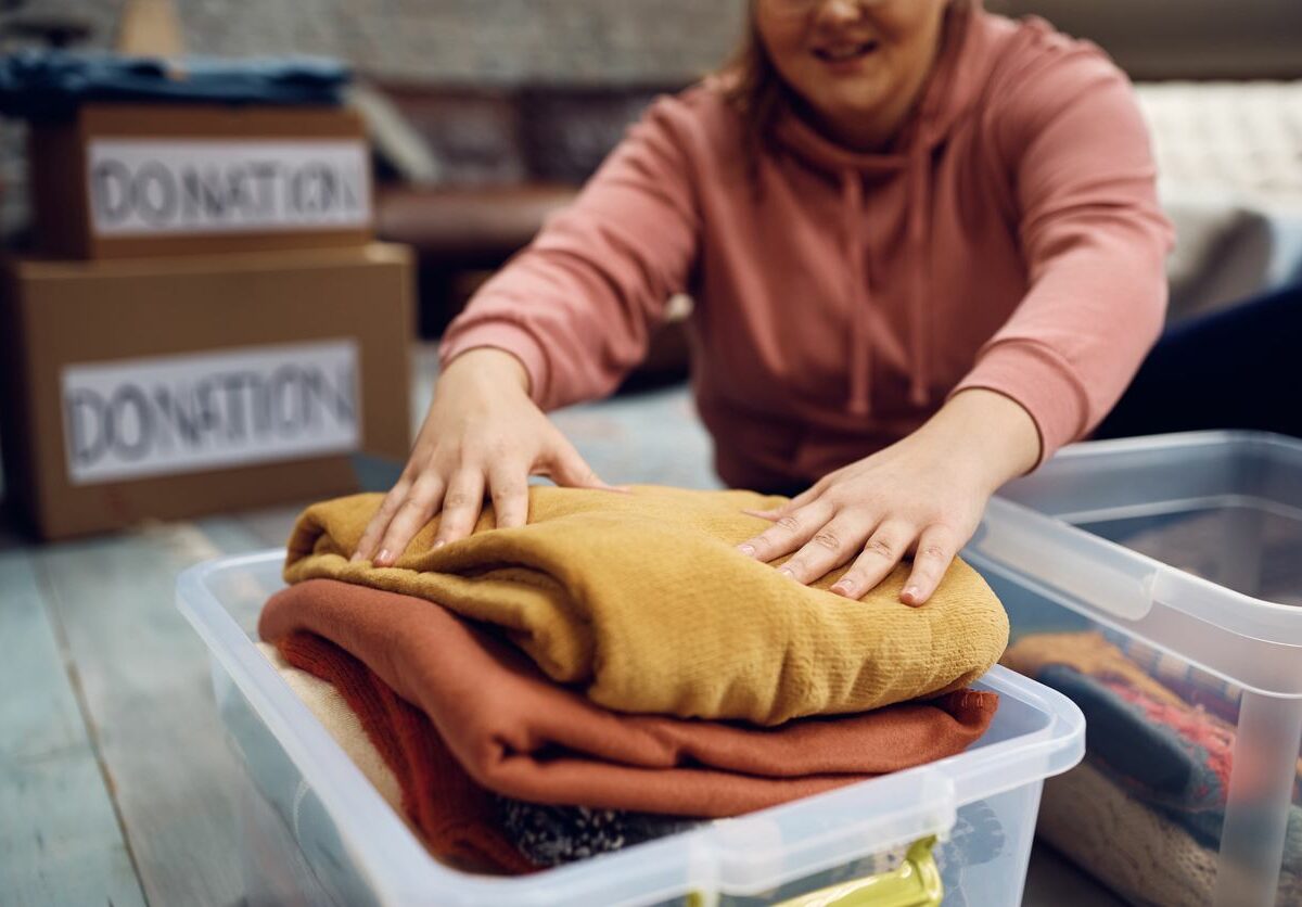 A woman stuffs folded clothing into a clear plastic bin with boxes labeled for donation in the background