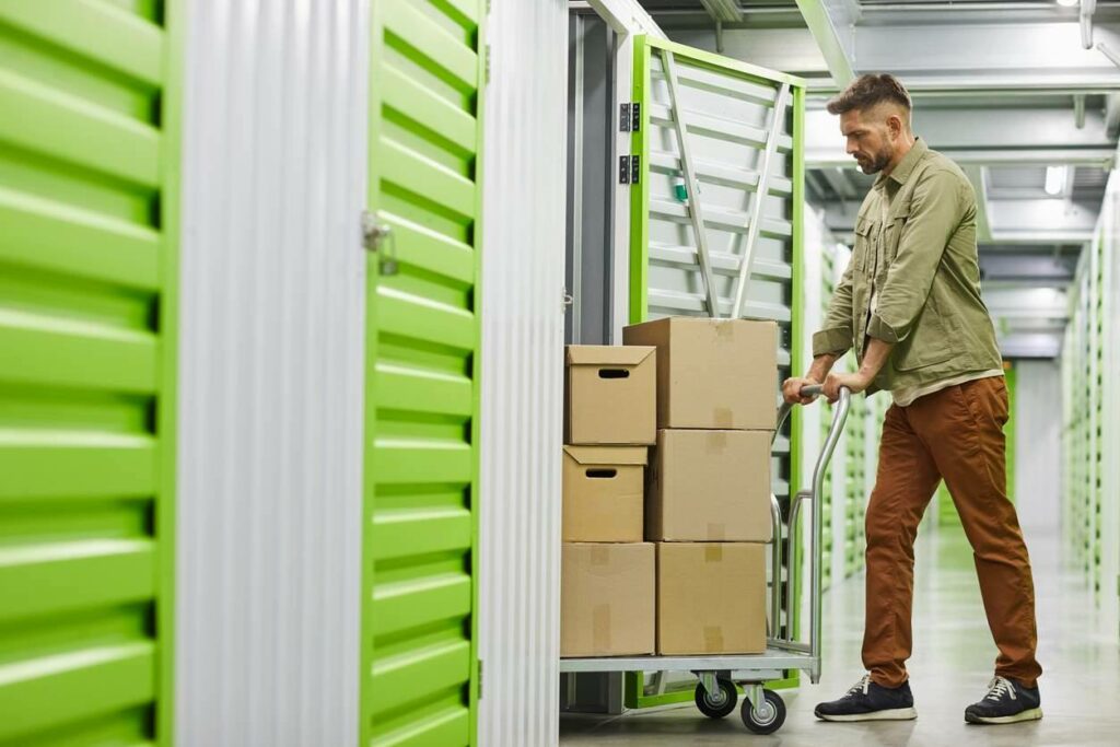 A man wheels a dolly with cardboard boxes into a storage unit. 