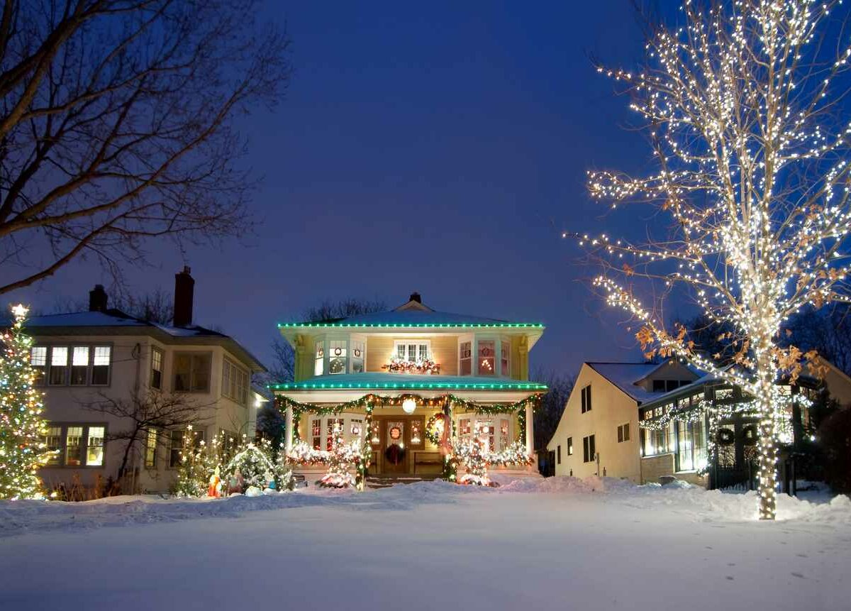 A home decorated in Christmas lights with green roof lights, white lights wrapped around trees, and glowing displays in a snowy yard