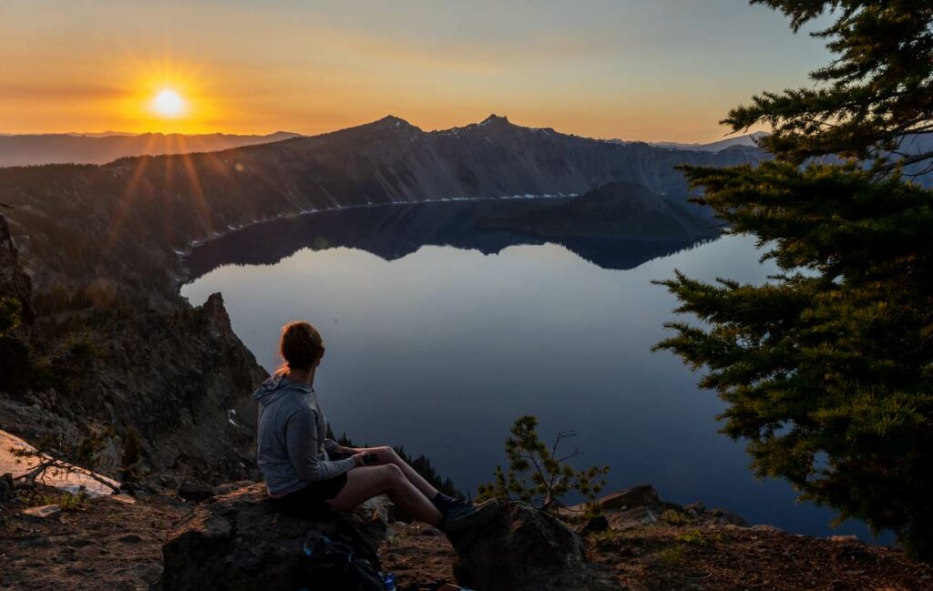 Woman with backpack on sits on a rock, watching the sunset over Crater Lake in summer